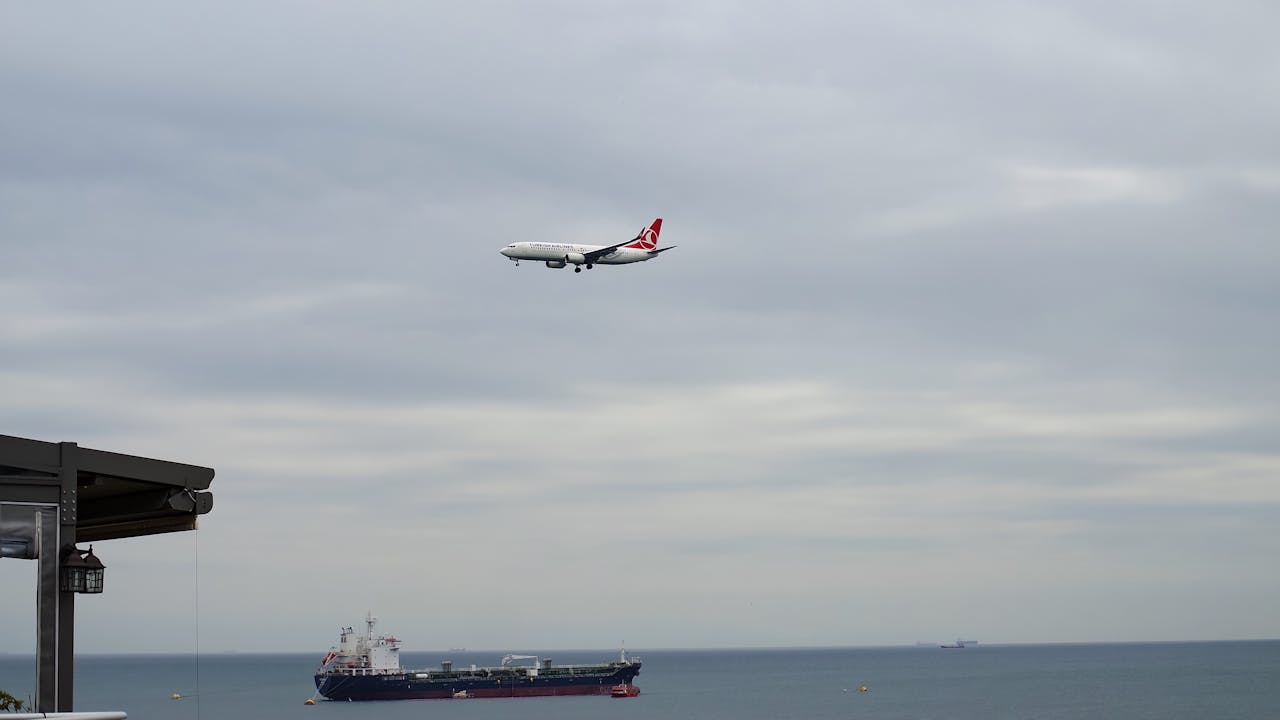 Airplane approaching over the sea with a cargo ship below under a cloudy sky.